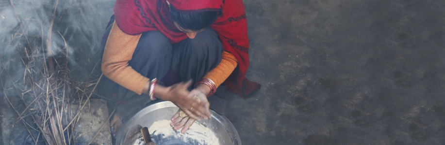 Woman squats over cooking equipment on ground with smoke swirling around